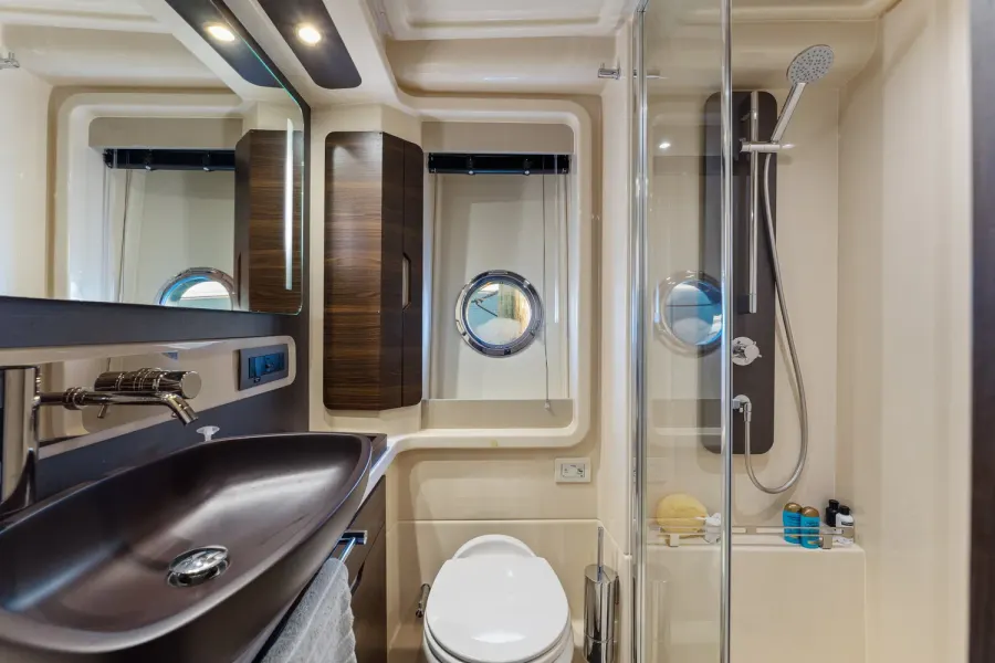 Compact yacht head with dark vessel sink, porthole, and glass shower